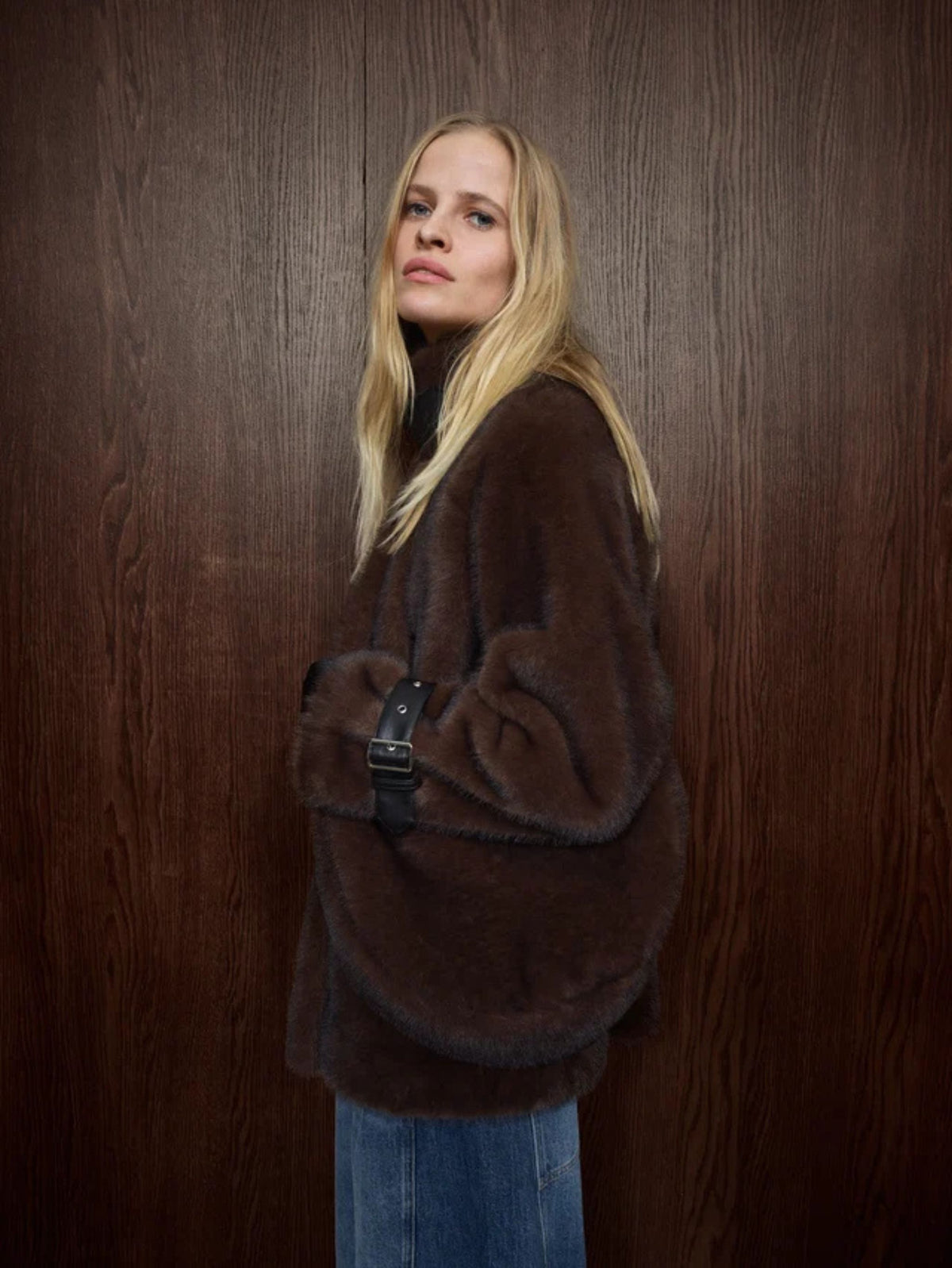 A blonde woman wearing a voluminous dark brown faux-fur jacket and blue jeans, holding a black shoulder bag with a silver buckle, posed against a dark brown wood-paneled wall.
