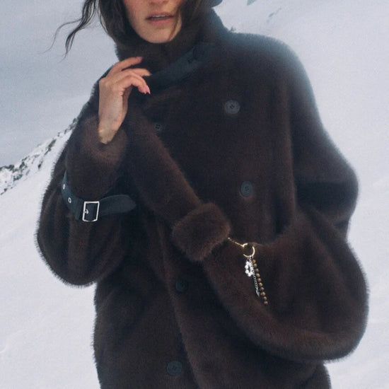 A woman wearing a dark brown fuzzy faux-fur coat with a high collar and large buttons, photographed outdoors against a snowy mountain background.