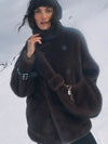A woman wearing a dark brown fuzzy faux-fur coat with a high collar and large buttons, photographed outdoors against a snowy mountain background.