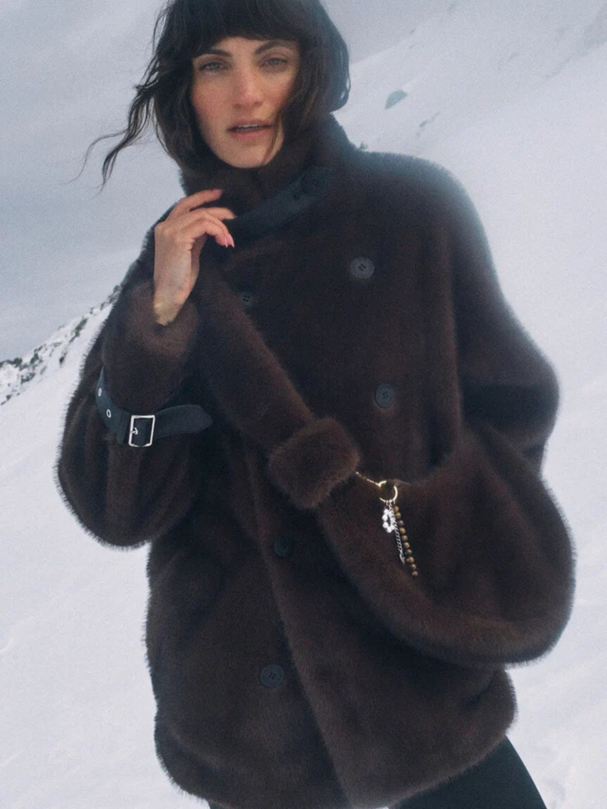 A woman wearing a dark brown fuzzy faux-fur coat with a high collar and large buttons, photographed outdoors against a snowy mountain background.