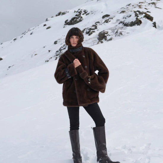 A person standing in a snowy mountain landscape wearing a dark brown plush hooded jacket, black pants, and tall black boots.