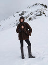 A person standing in a snowy mountain landscape wearing a dark brown plush hooded jacket, black pants, and tall black boots.