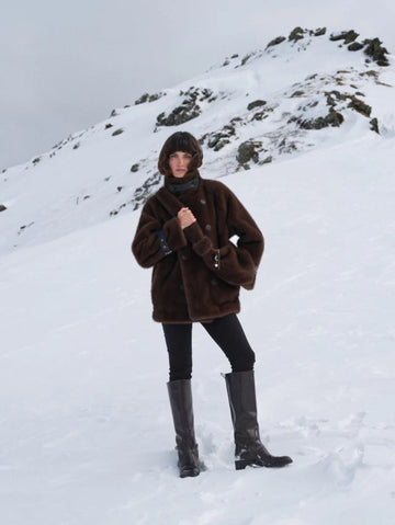 A person standing in a snowy mountain landscape wearing a dark brown plush hooded jacket, black pants, and tall black boots.