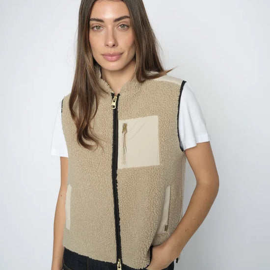 Woman wearing a beige fleece vest with a white shirt and black pants on a plain background