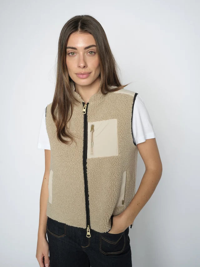 Woman wearing a beige fleece vest with a white shirt and black pants on a plain background