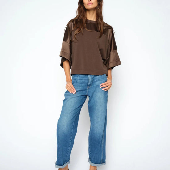 A woman modeling a brown short-sleeve top with sheer mesh sleeves and wide cuffs, paired with blue jeans and black slingback heels, against a plain white studio background.