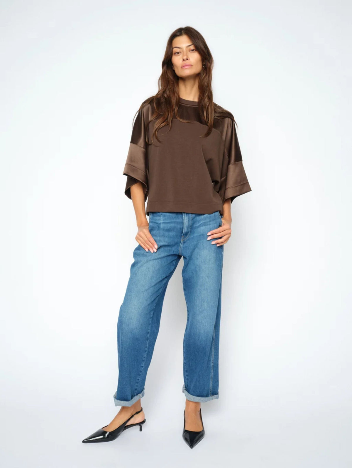 A woman modeling a brown short-sleeve top with sheer mesh sleeves and wide cuffs, paired with blue jeans and black slingback heels, against a plain white studio background.