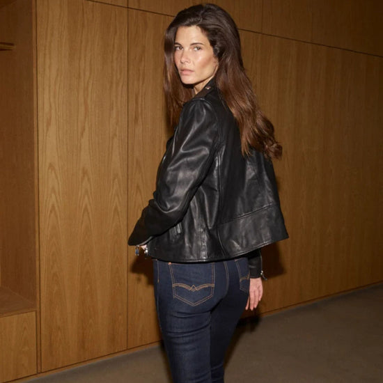 Woman wearing a black leather jacket and blue jeans against a wooden wall.