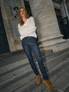 Woman standing on stone steps wearing a beige sweater, dark jeans, and brown boots.