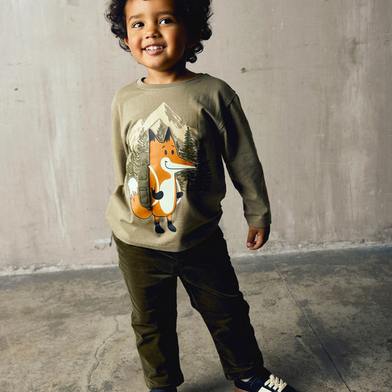A smiling young boy with curly dark hair stands in a studio setting wearing a beige long-sleeve crewneck sweatshirt with a graphic of an orange fox, dark olive pants, and navy sneakers.