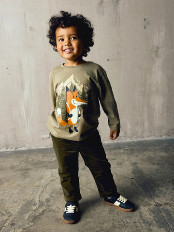 A smiling young boy with curly dark hair stands in a studio setting wearing a beige long-sleeve crewneck sweatshirt with a graphic of an orange fox, dark olive pants, and navy sneakers.