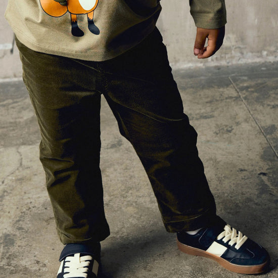 A child wearing dark olive green corduroy pants, a beige graphic sweatshirt, and navy blue sneakers with white side stripes and laces, standing on a concrete floor.