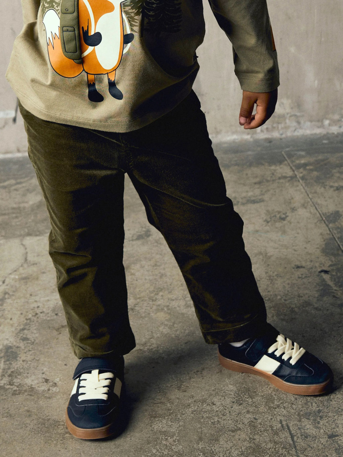 A child wearing dark olive green corduroy pants, a beige graphic sweatshirt, and navy blue sneakers with white side stripes and laces, standing on a concrete floor.