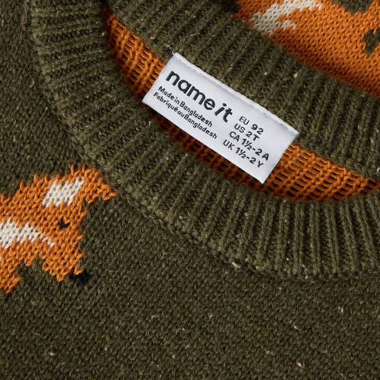 Close-up of a ribbed knit sweater neck showing a white 'name it' brand tag, with an olive green knit body and orange and white fox pattern.