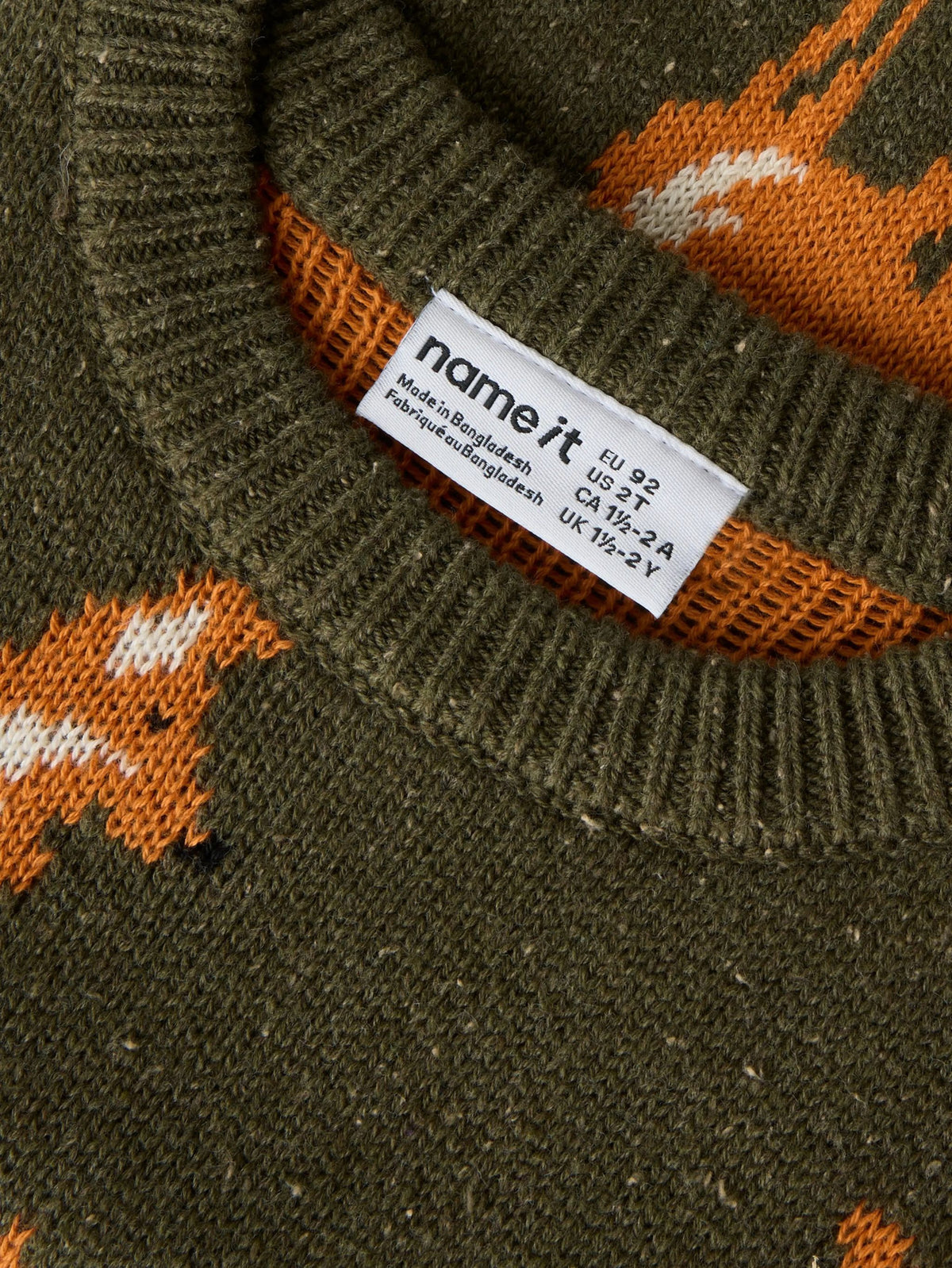 Close-up of a ribbed knit sweater neck showing a white 'name it' brand tag, with an olive green knit body and orange and white fox pattern.