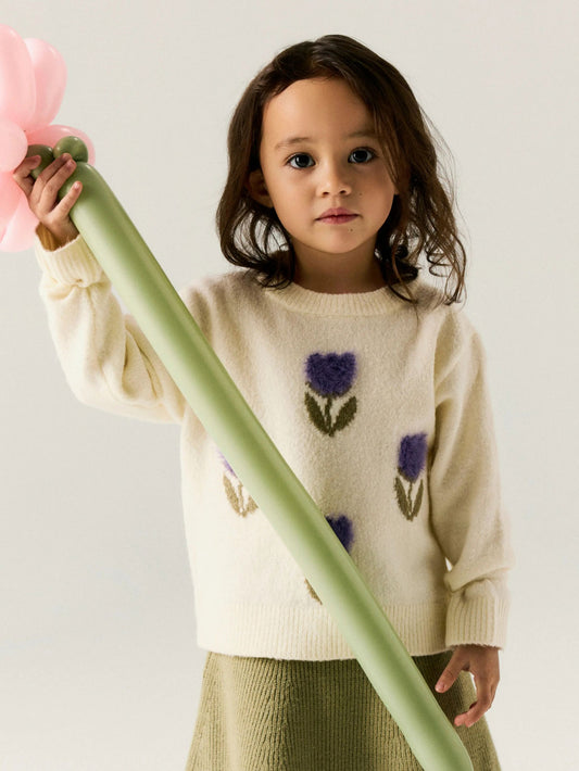 A young child wearing a cream knit sweater with purple tulip motifs and holding a large pink flower prop against a light gray studio background.