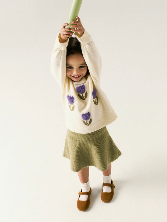 Child wearing a floral sweater and skirt holding a green object against a white background