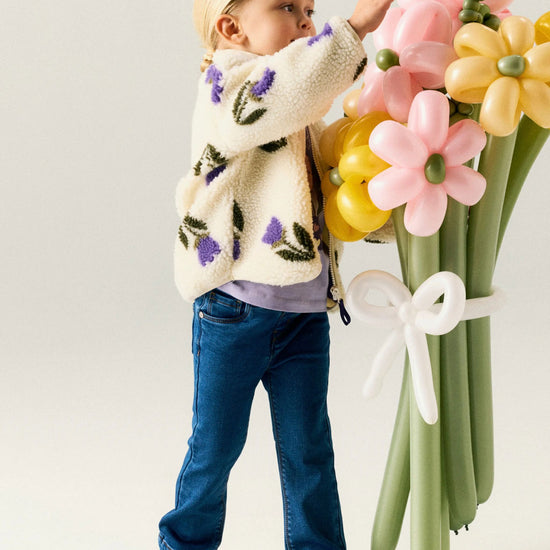 A young child stands beside a large balloon flower bouquet, wearing a cream sherpa cardigan with purple and green floral embroidery, blue jeans, and white sneakers against a plain light background.