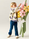 A young child stands beside a large balloon flower bouquet, wearing a cream sherpa cardigan with purple and green floral embroidery, blue jeans, and white sneakers against a plain light background.