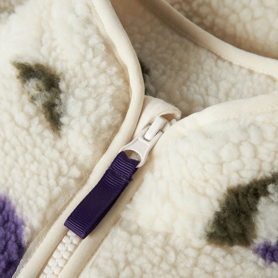 Close-up of a cream sherpa fleece zippered garment with a purple zipper pull tab and subtle dark and purple patterned accents.