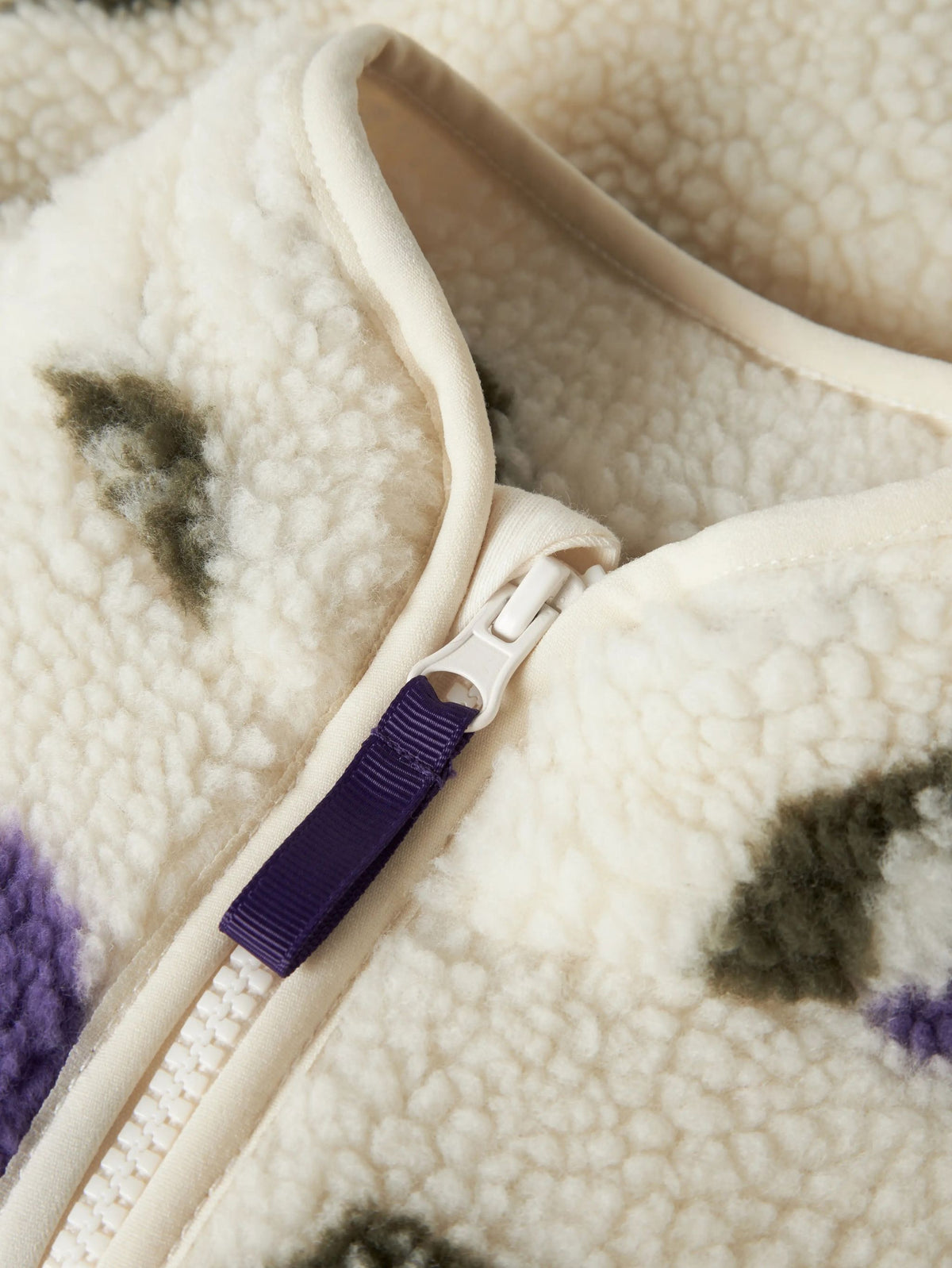 Close-up of a cream sherpa fleece zippered garment with a purple zipper pull tab and subtle dark and purple patterned accents.
