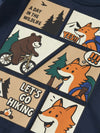 Close-up of a navy blue garment featuring a large cartoon-style multicolor graphic print with panels showing a bear on a bicycle, foxes, trees, campfire, and text reading 'A DAY IN THE WILDLIFE' and 'LET'S GO HIKING'.