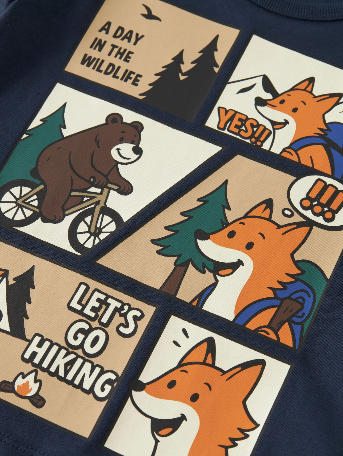Close-up of a navy blue garment featuring a large cartoon-style multicolor graphic print with panels showing a bear on a bicycle, foxes, trees, campfire, and text reading 'A DAY IN THE WILDLIFE' and 'LET'S GO HIKING'.