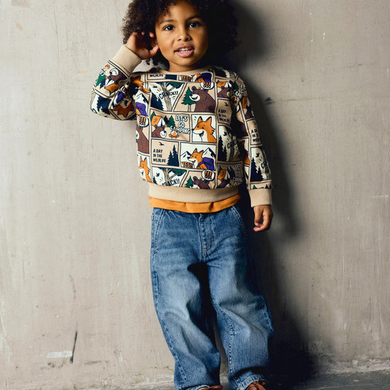 A young child with curly dark hair stands against a textured concrete wall wearing a long-sleeve crewneck sweatshirt with a colorful cartoon forest pattern, blue jeans, and brown leather shoes.