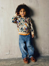 A young child with curly dark hair stands against a textured concrete wall wearing a long-sleeve crewneck sweatshirt with a colorful cartoon forest pattern, blue jeans, and brown leather shoes.