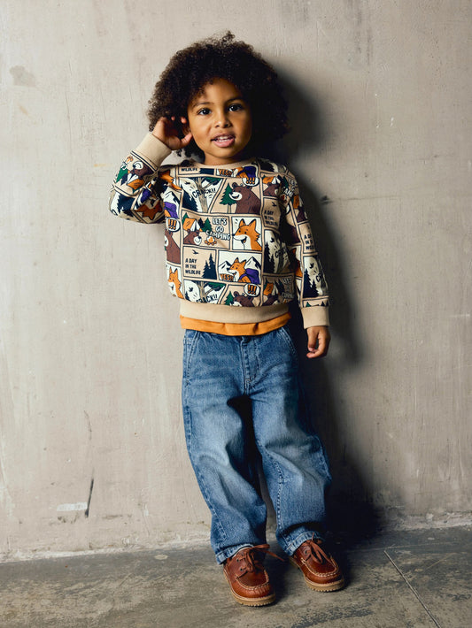 A young child with curly dark hair stands against a textured concrete wall wearing a long-sleeve crewneck sweatshirt with a colorful cartoon forest pattern, blue jeans, and brown leather shoes.