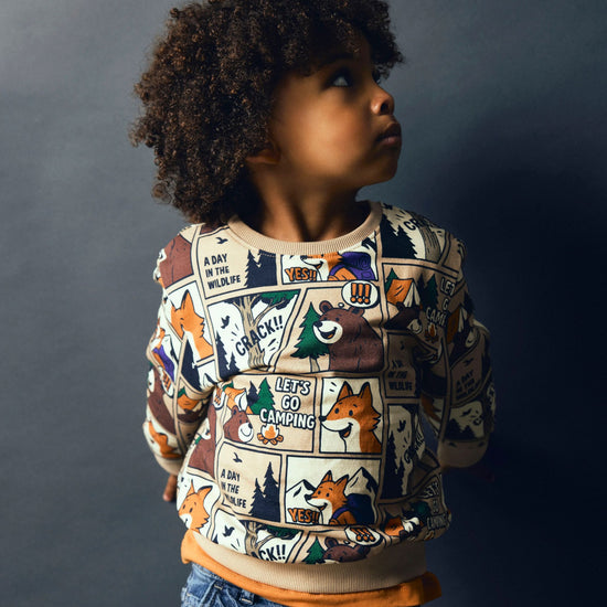 A child with curly dark hair wearing a long-sleeve crewneck sweatshirt with a detailed forest and campfire graphic print, paired with blue jeans, set against a dark gray studio background.