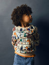 A child with curly dark hair wearing a long-sleeve crewneck sweatshirt with a detailed forest and campfire graphic print, paired with blue jeans, set against a dark gray studio background.