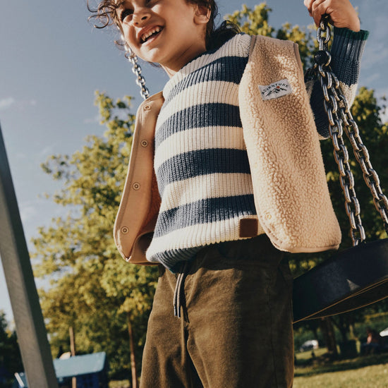 A child wearing a beige sherpa-lined jacket over a navy and cream striped sweater, standing on a swing at an outdoor playground.