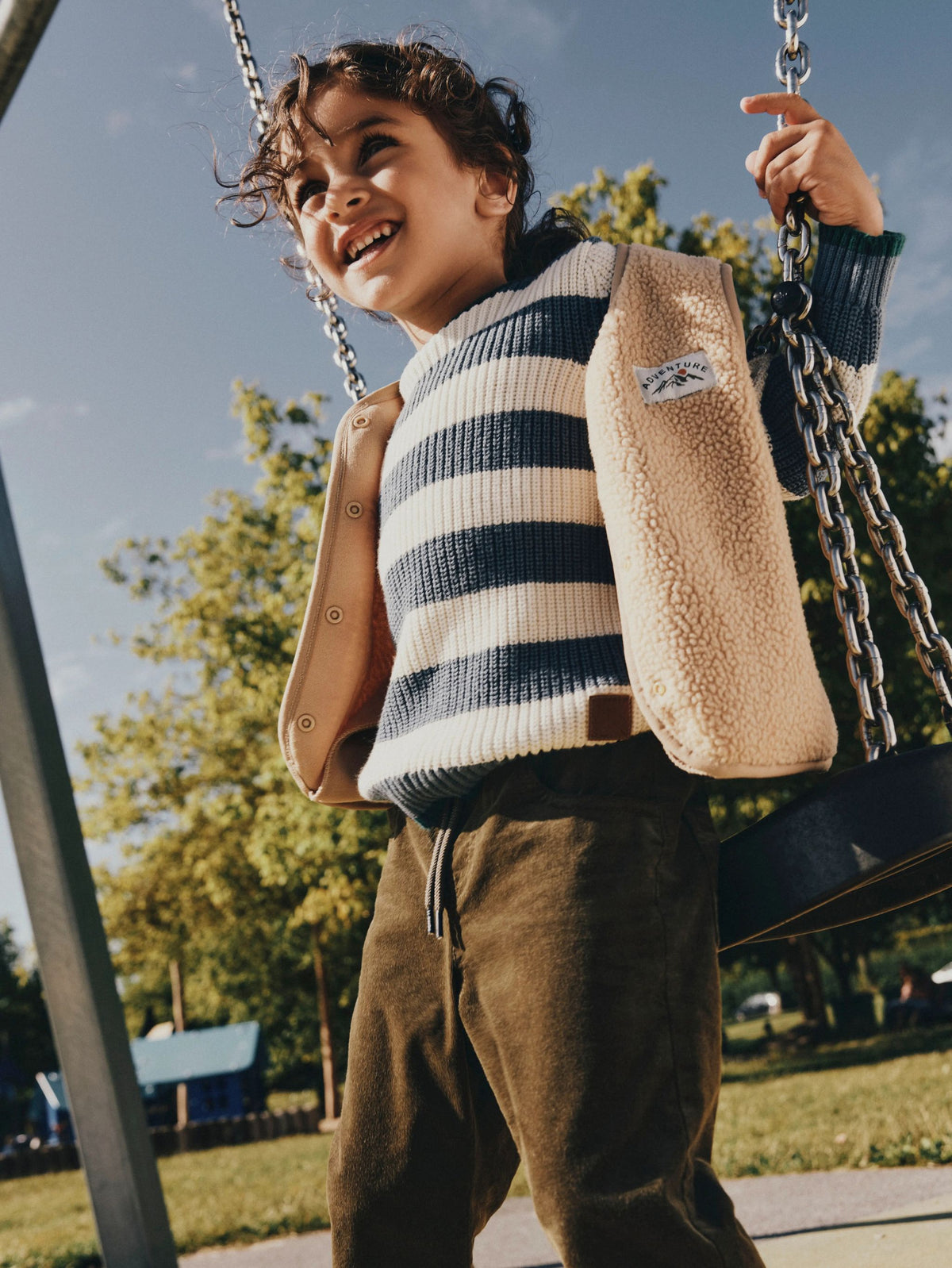 A child wearing a beige sherpa-lined jacket over a navy and cream striped sweater, standing on a swing at an outdoor playground.
