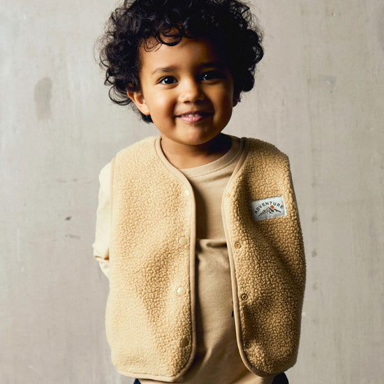 A boy standing in a beige sleeveless teddybear vest against a grey background