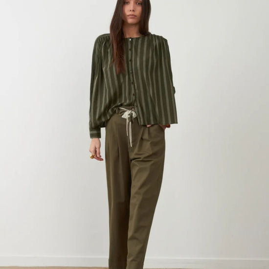 A woman stands against a plain off-white wall wearing an olive green vertically striped long-sleeve blouse with a drawstring waist, paired with olive green wide-leg trousers and dark brown lace-up boots.