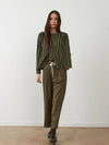 A woman stands against a plain off-white wall wearing an olive green vertically striped long-sleeve blouse with a drawstring waist, paired with olive green wide-leg trousers and dark brown lace-up boots.