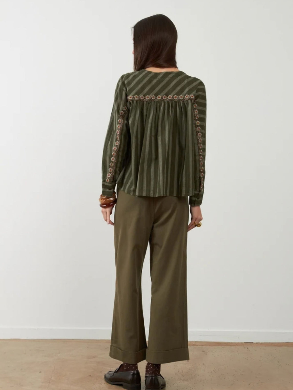 A woman is shown from the back wearing a green striped long-sleeve top with embroidered details down the sleeves and a pleated back, paired with olive green wide-leg pants and dark brown shoes, set against a plain white wall.