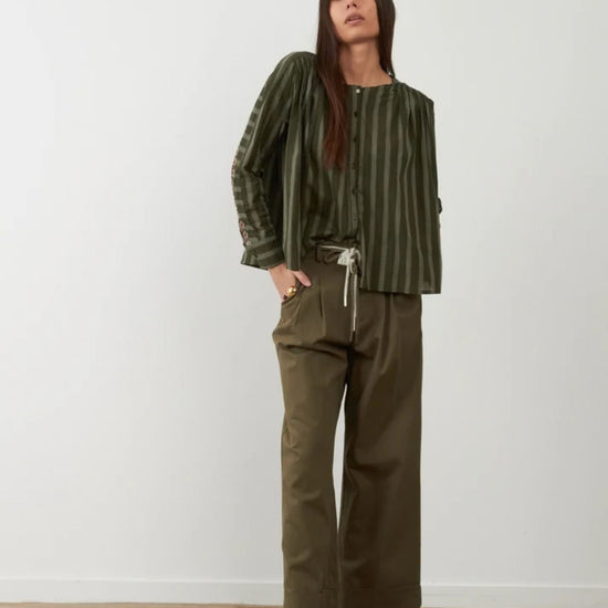 A woman stands against a plain white wall wearing an olive green vertically striped long-sleeve top with a drawstring waist, paired with matching olive green wide-leg cropped pants and dark brown lace-up shoes.
