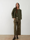 A woman stands against a plain white wall wearing an olive green vertically striped long-sleeve top with a drawstring waist, paired with matching olive green wide-leg cropped pants and dark brown lace-up shoes.