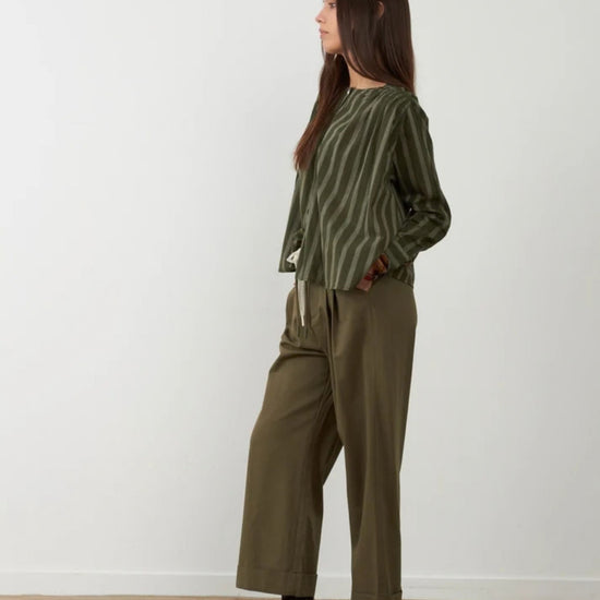A woman stands in profile wearing a green striped long-sleeve top and olive green wide-leg pants, paired with dark brown leather loafers, against a plain white wall and light wood floor.