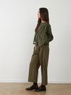 A woman stands in profile wearing a green striped long-sleeve top and olive green wide-leg pants, paired with dark brown leather loafers, against a plain white wall and light wood floor.
