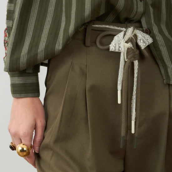 Close-up of a person wearing olive green striped long-sleeve top and matching olive green pleated trousers, with a lace-trimmed drawstring belt and a gold ring on the hand.
