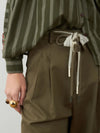 Close-up of a person wearing olive green striped long-sleeve top and matching olive green pleated trousers, with a lace-trimmed drawstring belt and a gold ring on the hand.