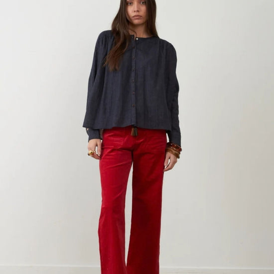 A woman stands against a plain off-white wall wearing a dark navy long-sleeve button-up top and bright red wide-leg pants, with black shoes and bracelets.