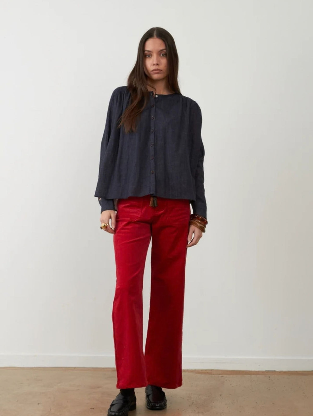 A woman stands against a plain off-white wall wearing a dark navy long-sleeve button-up top and bright red wide-leg pants, with black shoes and bracelets.