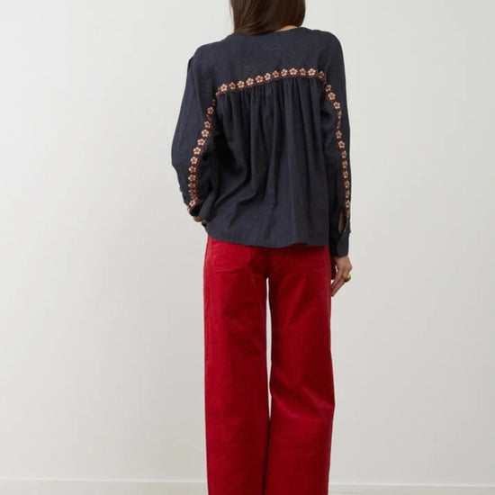 A woman viewed from behind wearing a dark navy long-sleeve blouse with orange floral embroidery down the back and red wide-leg pants, standing against a plain white wall.
