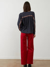A woman viewed from behind wearing a dark navy long-sleeve blouse with orange floral embroidery down the back and red wide-leg pants, standing against a plain white wall.
