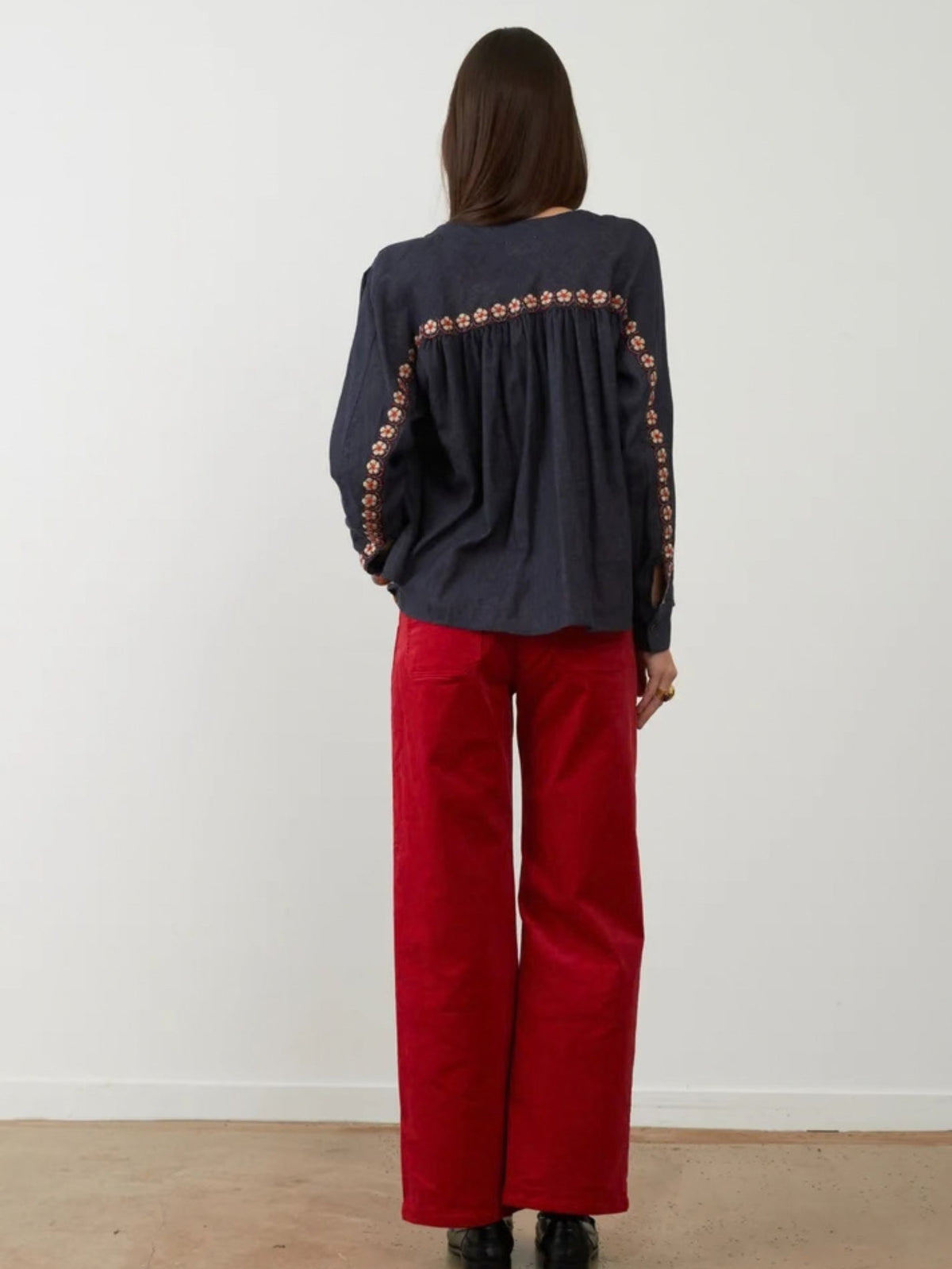 A woman viewed from behind wearing a dark navy long-sleeve blouse with orange floral embroidery down the back and red wide-leg pants, standing against a plain white wall.