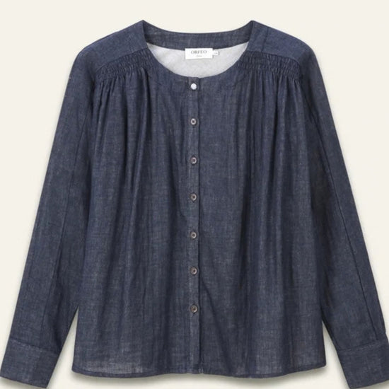 A dark blue chambray button-up blouse with a smocked neckline, long sleeves, and a relaxed fit, shown against a light cream background.